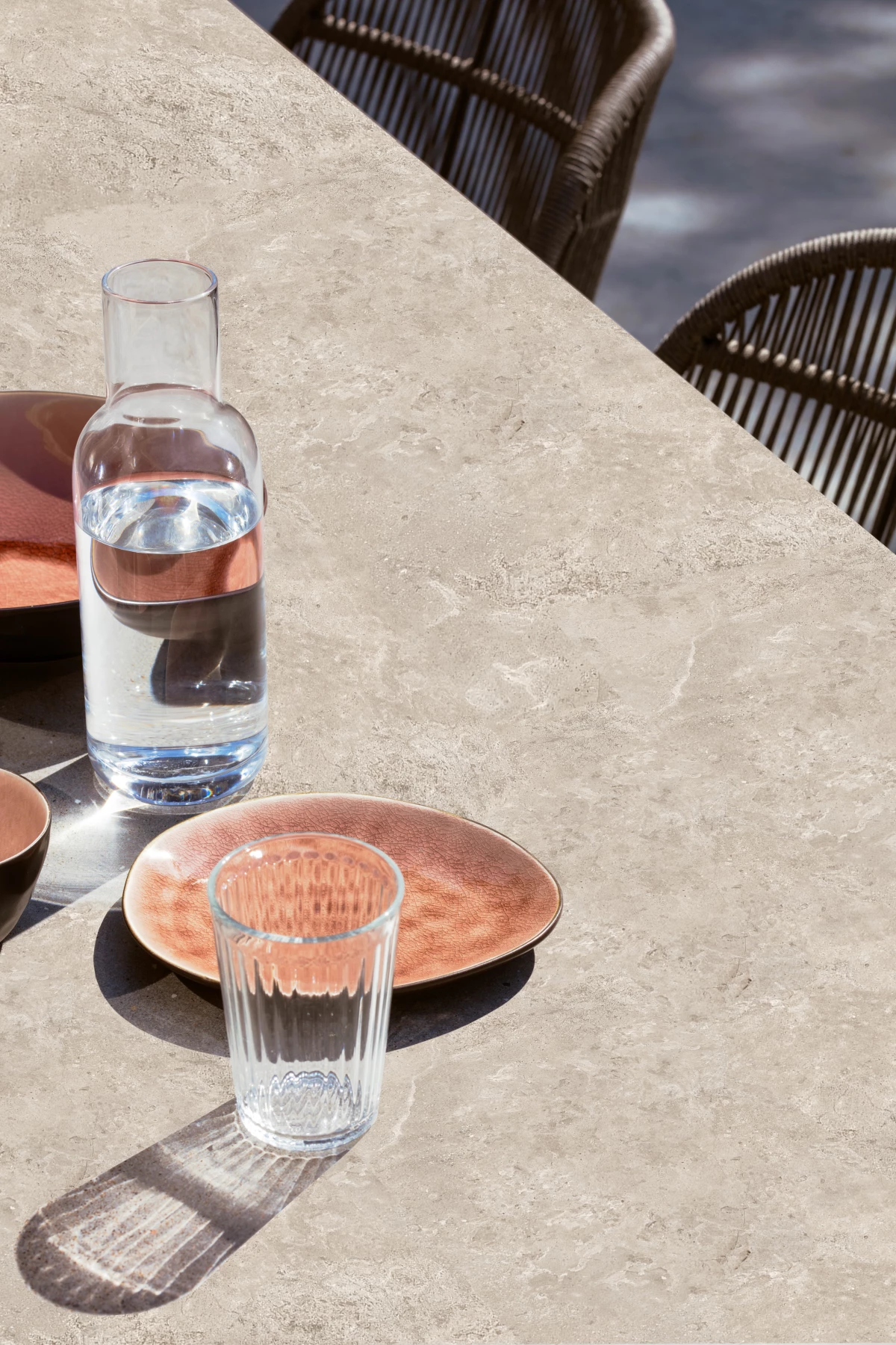 Close-up of the Kodo Dining Table by Vincent Sheppard, featuring a Sabbia Panna ceramic stone top, styled with glassware and copper-toned plates, with woven dining chairs in the background.